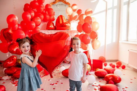Young boy and girl smiling while holding a large red heart shaped balloon, celebrating love and friendship surrounded by scattered red confetti and many balloonsの写真素材