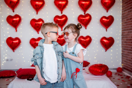 Joyful boy and girl wearing sunglasses, sharing a tender moment surrounded by red heart-shaped balloons and lights, symbolizing young love and friendship on Valentine's Dayの写真素材
