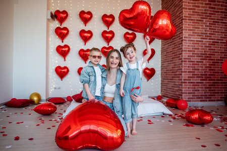 Mother and her two children posing, smiling, and celebrating Valentine's Day, surrounded by numerous red heart-shaped balloons and scattered rose petals, symbolizing family love and joyの写真素材