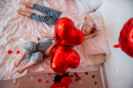 Two young children relaxing on a bed decorated with red heart balloons and rose petals, celebrating love and childhood happiness during Valentines Dayの写真素材