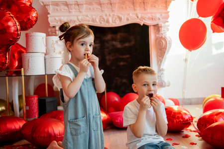 Little girl and boy enjoying French fries on the floor surrounded by red heart-shaped and regular balloons, celebrating a special occasion with love and joyの写真素材
