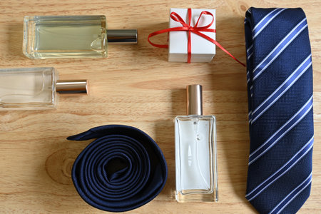 Flatlay of father's day gifts, square white gift box with red bow and tag with "happy father's day" message, blue striped tie, three square perfumes, on pine wood background with copy spaceの写真素材