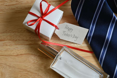 Flatlay of father's day gifts, square white gift box with red bow and tag with "happy father's day" message, blue striped tie, square perfume, on pine wood background with copy spaceの写真素材