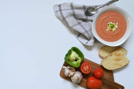 Flatlay of table with white plate with Spanish cordovan salmorejo gazpacho next to dark wood table with tomato, green pepper, garlic and toast, on a checkered tablecloth and a white surface, with copy spaceの写真素材