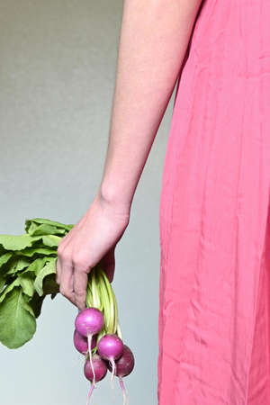 Close-up of young latin woman's hand wearing pink dress and holding bunch of ripe radishes in her hand, gray background and copy spaceの写真素材