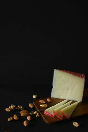 Vertical closeup of manchego cheese next to nuts, cutting board, black background and copy spaceの写真素材