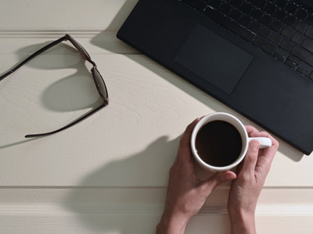 Overhead shot of woman's hands with white cup with coffee next to sunglasses and laptop, on white wooden desk, strong shadows, copy spaceの写真素材
