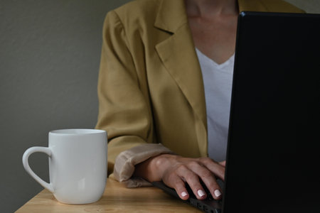 Close-up of young latin woman with jacket working with a laptop next to cup of coffee on wooden...