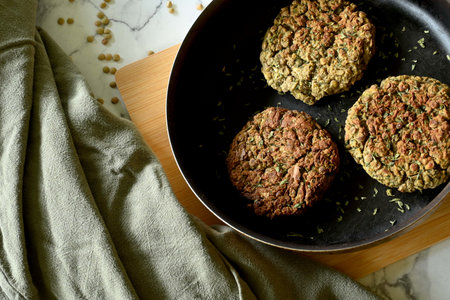 Overhead shot of lentil burgers in frying pan, on cutting board, next to cloth, on marble table