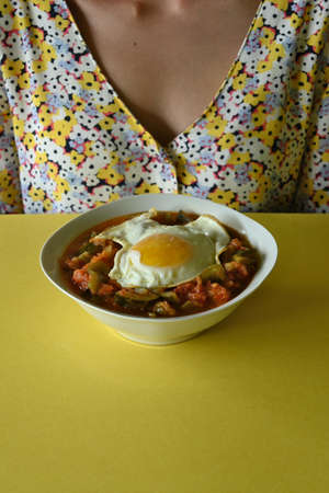 Selective focus of pisto dish with fried egg on yellow table, woman in backgroundの写真素材