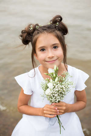 a child girl holds a bouquet of white flowers on a blurry background.dark hair , white flowers in her hands, white dressの写真素材