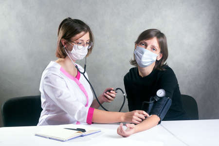 a young woman doctor in a medical gown and mask measures blood pressure with a tonometer to a masked womanの写真素材