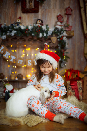 Happy little girl in pajamas playing with bunny near a fireplace at Christmas Eveの写真素材