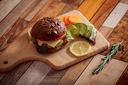 Dark burger with grain bread on dark ceramic plate, salad, rosemary and lemonの写真素材