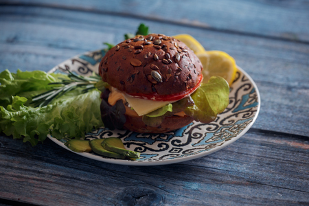Dark burger with grain bread on dark ceramic plate, salad, rosemary and lemonの写真素材