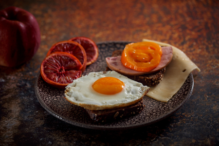 Grain bread with fried egg, tomato, cheese and bacon on ceramic plate with red orange to desertの写真素材