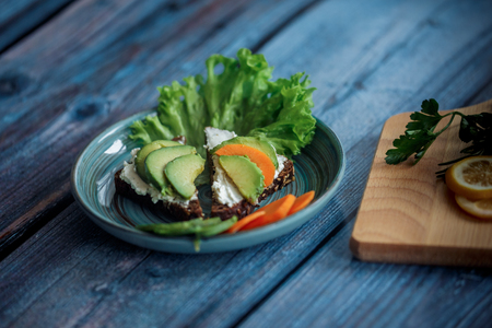 Dark burger with grain bread on dark ceramic plate, salad, rosemary and lemonの写真素材
