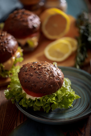Dark burger with grain bread on dark ceramic plate, salad, rosemary and lemonの写真素材