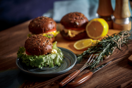 Dark burger with grain bread on dark ceramic plate, salad, rosemary and lemonの写真素材