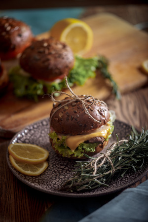 Dark burger with grain bread on dark ceramic plate, salad, rosemary and lemonの写真素材