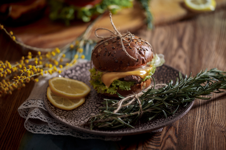 Dark burger with grain bread on dark ceramic plate, salad, rosemary and lemonの写真素材