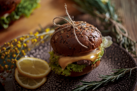 Dark burger with grain bread on dark ceramic plate, salad, rosemary and lemonの写真素材