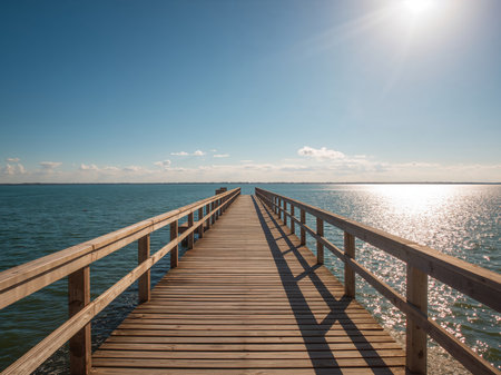 A captivating view of a wooden pier extending into a serene sea. The bright sunlight reflects on the water, creating a shimmering effect. The clear blue sky adds to the peaceful atmosphere.の素材