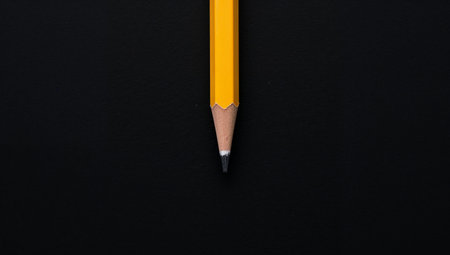 A bright yellow, hexagonal pencil rests against a stark black background The tip is sharpened, ready for use The composition is simple, focused and direct.の素材