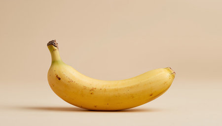 A single banana, predominantly yellow with brown spots, curves against a plain, light beige background. Soft, diffused lighting creates a subtle shadow.の素材