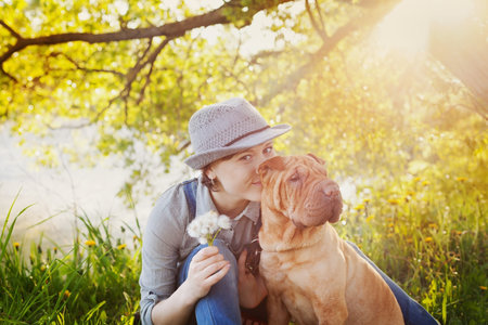 happy young woman in denim overalls and hat with a bouquet of dandelions kissing her red cute dog...