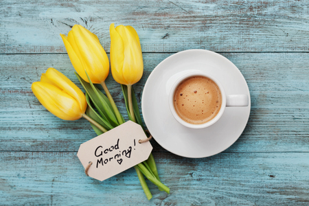 Coffee mug with yellow tulip flowers and notes good morning on blue rustic table from above, breakfast on Mothers day or Womens day