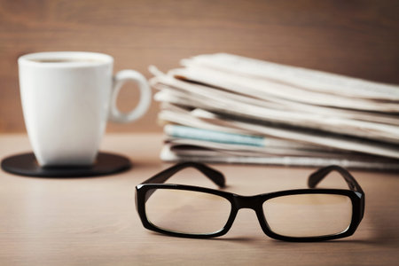 Eyeglasses, coffee mug and stack of newspapers on wooden desk for themes of ophthalmology, poor...