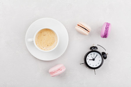 Morning cup of coffee, cake macaron and alarm clock on light table top view in flat lay style....