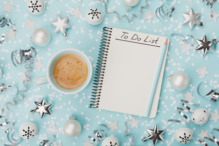 Cup of coffee and notebook with to do list decorated with confetti, silver stars and balls on blue background top view. Christmas planning concept. Flat lay.の写真素材
