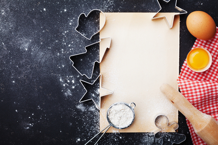 Food background for christmas baking. Cookies cutters, flour, rolling pin, eggs and paper sheet on kitchen table top view. の写真素材