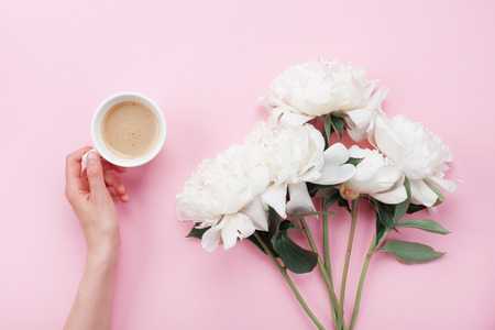 Woman hand with cup of coffee and beautiful white peony flowers on pink pastel table top view. Cozy breakfast in flat lay style.の写真素材