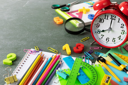 Back to school and education concept with red alarm clock and colorful school supplies on blackboard...