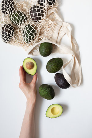 Woman hand holding half avocado and whole avocados in bag on table top view. Eco friendly concept, flat lay.の写真素材
