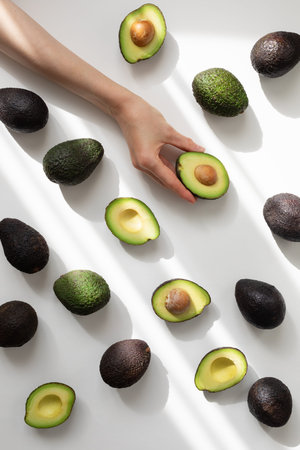 Woman hand holding half avocado of heap whole avocados on table top view in aesthetic shadow...