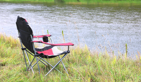 Portable folding tourist red armchair on green grass in summer on picnic near river