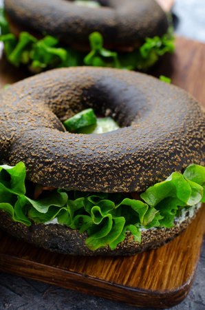Black Bagels Sandwiches with Cucumbers, Salad and Ham on Wooden Board and Dark Background, Tasty Snack or Breakfastの写真素材