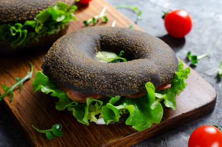 Black Bagels Sandwiches with Cucumbers, Salad and Ham on Wooden Board and Dark Background, Tasty Snack or Breakfastの写真素材
