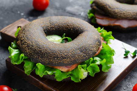 Black Bagels Sandwiches with Cucumbers, Salad and Ham on Wooden Board and Dark Background, Tasty Snack or Breakfastの写真素材