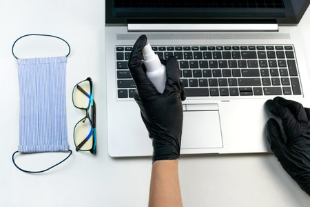Two women's hands in black latex gloves disinfect a computer with an antibacterial spray. Office...