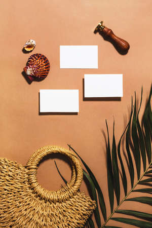 Empty stationery minimal mockup template. Paper business card, woven straw bag, tropical sea decor, palm tree leaf, sealing wax stamp. Summer trendy flat lay on warm ochre background. Creative conceptの写真素材