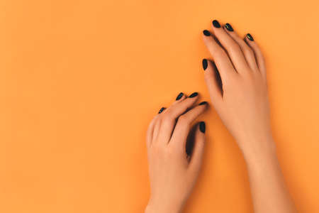 Two tender womans hands on Marigold orange background. Perfect black matte manicure and holographic metallic design. Beauty spa salon flat lay banner concept, copy space. Trendy color nail gel polishの写真素材