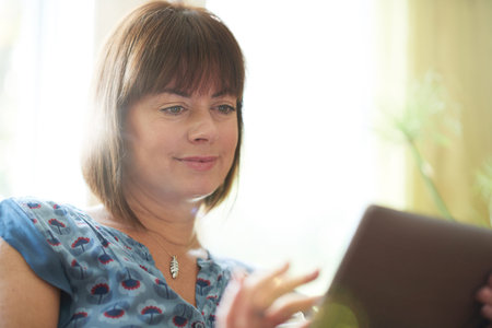Serene lady in her forties reading emails on a wireless tablet in a brightly lit house- filtered...