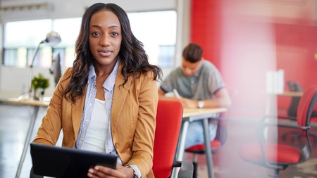 Casual portrait of an african american business woman using technology in a bright and sunny startup with the team in the backgroundの写真素材