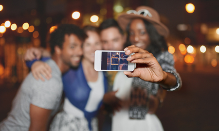 Multi-ethnic millenial group of friends taking a selfie photo with mobile phone on rooftop terrasse using flash at night timeの写真素材