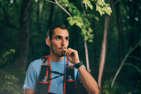 Fit male jogger hydrates while day training for cross country forest trail race in a nature park.